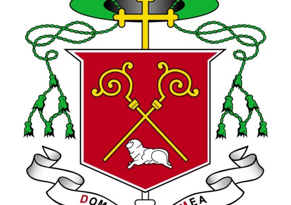 Coat of arms featuring a red shield with two golden crosiers crossed behind a lamb, topped with a green hat and a gold cross. Decorative green tassels hang from the hat, with a ribbon below displaying the Latin phrase "Dominus Spes Mea."