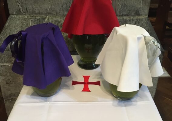Three decorative vases covered with colored cloths—purple, red, and white—arranged on a white tablecloth featuring a red cross, set in a church interior.