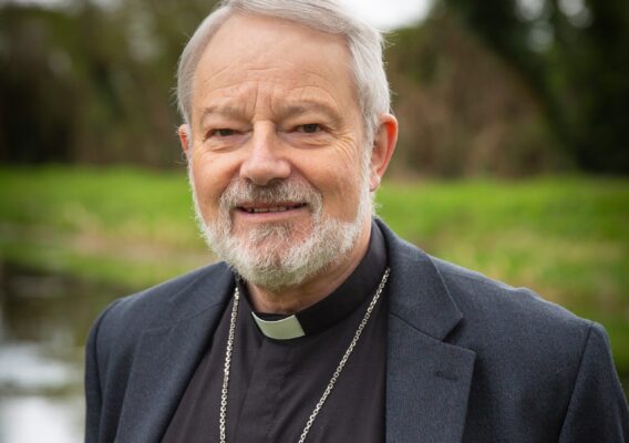 Portrait of Bishop Dornan with a beard, wearing a clerical collar and a dark jacket, standing outdoors near a body of water, with greenery in the background.