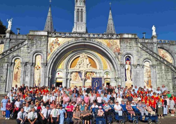 Group photo of visitors at the Basilica of Our Lady of Lourdes, featuring a large crowd in front of the ornate stone facade and colorful murals, with clear blue skies in the background.