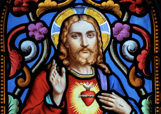 Stained glass depiction of Jesus Christ with a radiant heart, surrounded by intricate floral patterns and vibrant colors, featuring the text "Dilexit Nos" at the top.