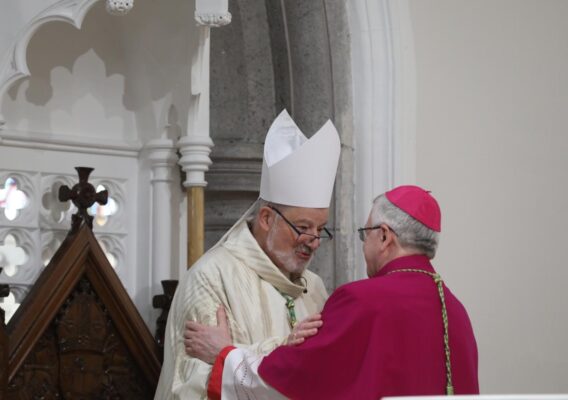 Two bishops in ceremonial attire engage in a warm greeting inside a church, showcasing the significance of religious fellowship. The bishop on the left wears a white mitre and ornate vestments, while the bishop on the right dons a pink mitre and robe, emphasizing their roles in the clergy.