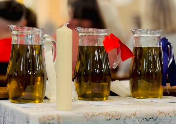 Three glass pitchers filled with a golden liquid are displayed on a lace tablecloth, accompanied by a white candle. The pitchers are adorned with colorful ribbons: red, blue, and white. The background features a blurred audience, suggesting a ceremonial or religious setting.