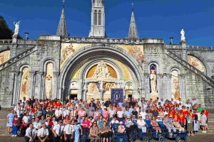 Group of pilgrims gathered in front of the Basilica of Our Lady of Lourdes, showcasing diverse attendees in colorful attire against a backdrop of historical architecture and clear blue sky.