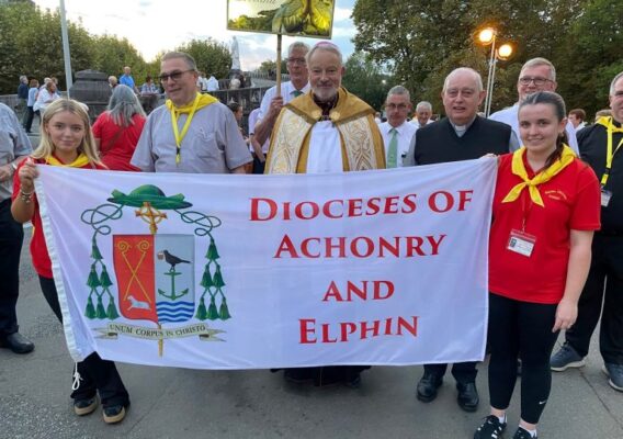 Group of participants holding a banner that reads "Dioceses of Achonry and Elphin" at a religious event, featuring clergy and attendees in traditional attire, with a scenic background.